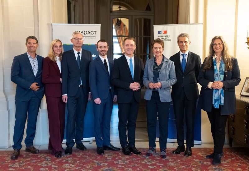 Eine Gruppe von acht Personen steht lächelnd in einem eleganten Raum, gekleidet in formellen Anzügen. Im Hintergrund sind zwei Banner zu sehen, eines mit der Aufschrift 'Ambassade de France en Autriche'. Der Boden ist mit einem roten Teppich bedeckt,